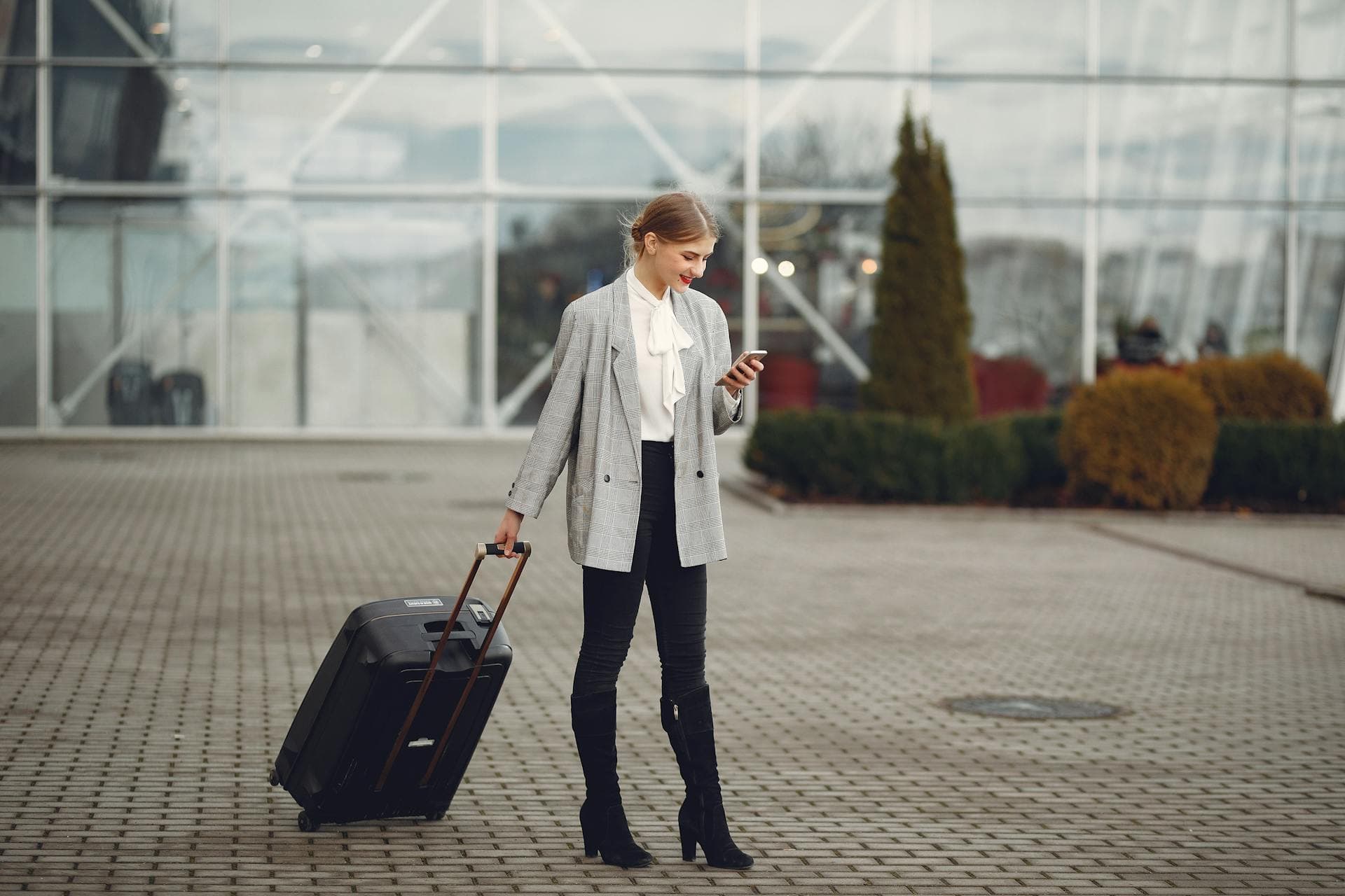 Smiling woman with suitcase checking phone outside airport